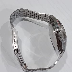 Puede incluir: Un reloj de pulsera de metal plateado con una correa de eslabones m&uacute;ltiples y un cierre desplegable. La caja del reloj es redonda con dos pulsadores laterales y una corona central, con un acabado met&aacute;lico y una tapa trasera grabada.