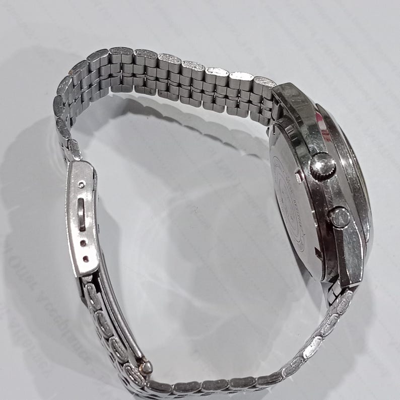 Puede incluir: Un reloj de pulsera de metal plateado con una correa de eslabones m&uacute;ltiples y un cierre desplegable. La caja del reloj es redonda con dos pulsadores laterales y una corona central, con un acabado met&aacute;lico y una tapa trasera grabada.