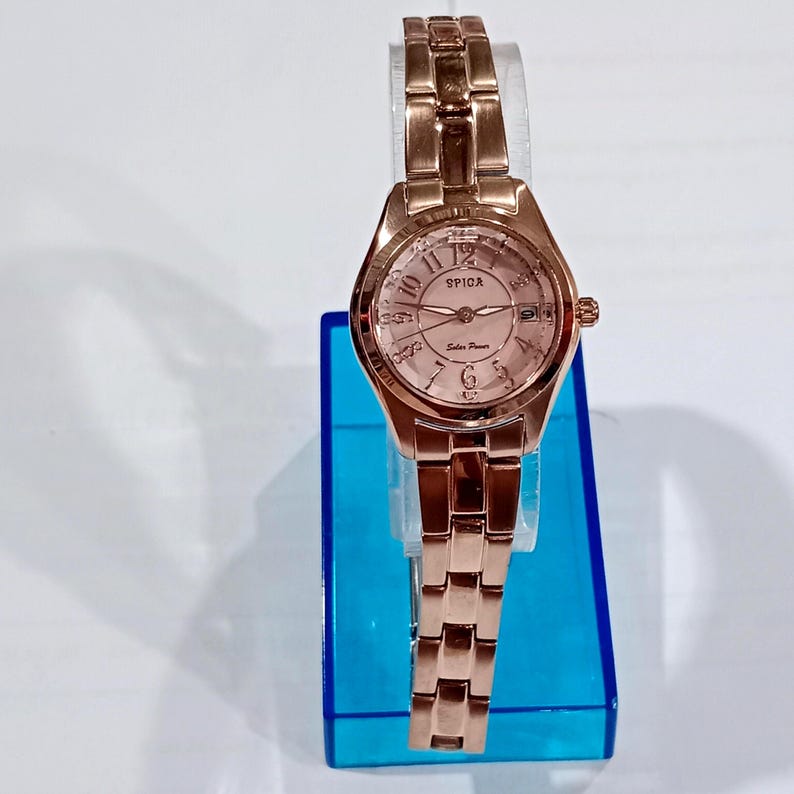 Puede incluir: Un reloj de pulsera Spica de energ&iacute;a solar en oro rosa. El reloj tiene una esfera redonda con una esfera rosa claro, agujas doradas y una correa de oro rosa a juego. El reloj se muestra sobre un soporte azul.