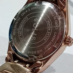 Puede incluir: Primer plano de un reloj de pulsera de oro rosa. La parte trasera del reloj es visible, mostrando texto que incluye "STEEL", "JAPAN MOV'T" y "SOLAR POWER". El reloj est&aacute; marcado "W.R. 10BAR".
