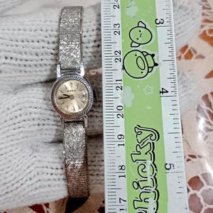 Op de afbeelding: Een vintage Rado polshorloge met een zilverkleurige, getextureerde band en een ronde wijzerplaat. De wijzerplaat is lichtgoudkleurig met zilveren wijzers en markeringen. Het horloge ligt naast een liniaal.