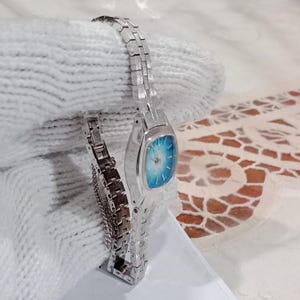 K&ouml;nnte beinhalten: Eine silberfarbene Armbanduhr mit rechteckigem Zifferblatt und hellblauem Farbverlauf. Die Uhr hat ein Metallarmband mit Gliedern. Die Uhr wird auf einer wei&szlig;en Oberfl&auml;che pr&auml;sentiert.
