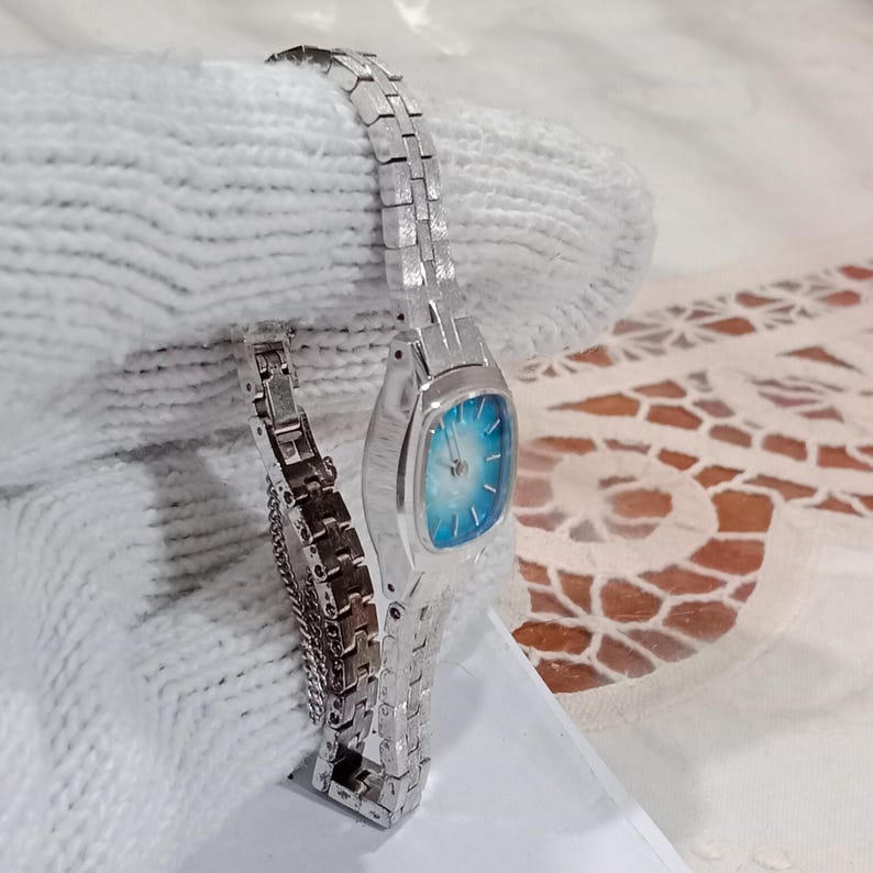 K&ouml;nnte beinhalten: Eine silberfarbene Armbanduhr mit rechteckigem Zifferblatt und hellblauem Farbverlauf. Die Uhr hat ein Metallarmband mit Gliedern. Die Uhr wird auf einer wei&szlig;en Oberfl&auml;che pr&auml;sentiert.