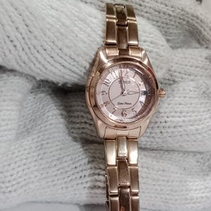 Puede incluir: Un reloj de pulsera solar de oro rosa con esfera rosa claro. El reloj tiene una correa met&aacute;lica y una esfera redonda con los n&uacute;meros del 1 al 12. La esfera del reloj tiene el texto "S.G.A." y "Solar Power".