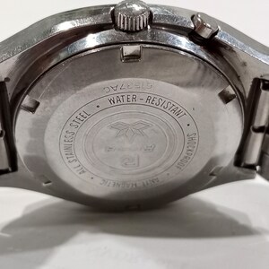 Puede incluir: La parte posterior de una caja de reloj de acero inoxidable en tono plateado con un dise&ntilde;o circular y texto grabado que incluye Water-Resistant, Shockproof, Anti-Magnetic y All Stainless Steel. El n&uacute;mero de serie 61537AC es visible arriba.