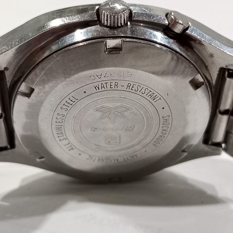 Puede incluir: La parte posterior de una caja de reloj de acero inoxidable en tono plateado con un dise&ntilde;o circular y texto grabado que incluye Water-Resistant, Shockproof, Anti-Magnetic y All Stainless Steel. El n&uacute;mero de serie 61537AC es visible arriba.
