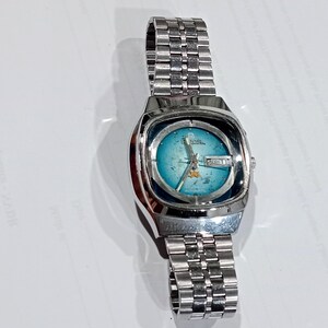 Puede incluir: Un reloj de pulsera plateado con correa de eslabones met&aacute;licos y caja rectangular redondeada. La esfera presenta un degradado azul verdoso con textura desgastada, manecillas plateadas y una ventana de d&iacute;a y fecha a las tres.