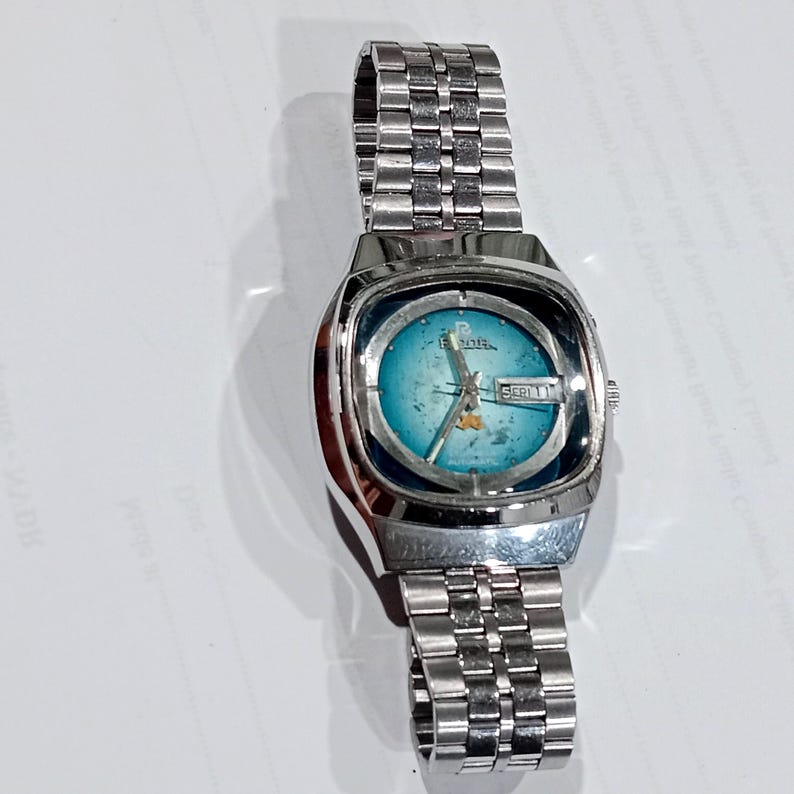 Puede incluir: Un reloj de pulsera plateado con correa de eslabones met&aacute;licos y caja rectangular redondeada. La esfera presenta un degradado azul verdoso con textura desgastada, manecillas plateadas y una ventana de d&iacute;a y fecha a las tres.