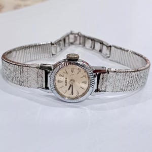 Op de afbeelding: Een vintage Rado polshorloge met een zilverkleurige, getextureerde band en een ronde wijzerplaat. De wijzerplaat is cr&egrave;mekleurig met zwarte wijzers en uurmarkeringen. Het horloge heeft een kleine, getextureerde kroon en een sluiting.