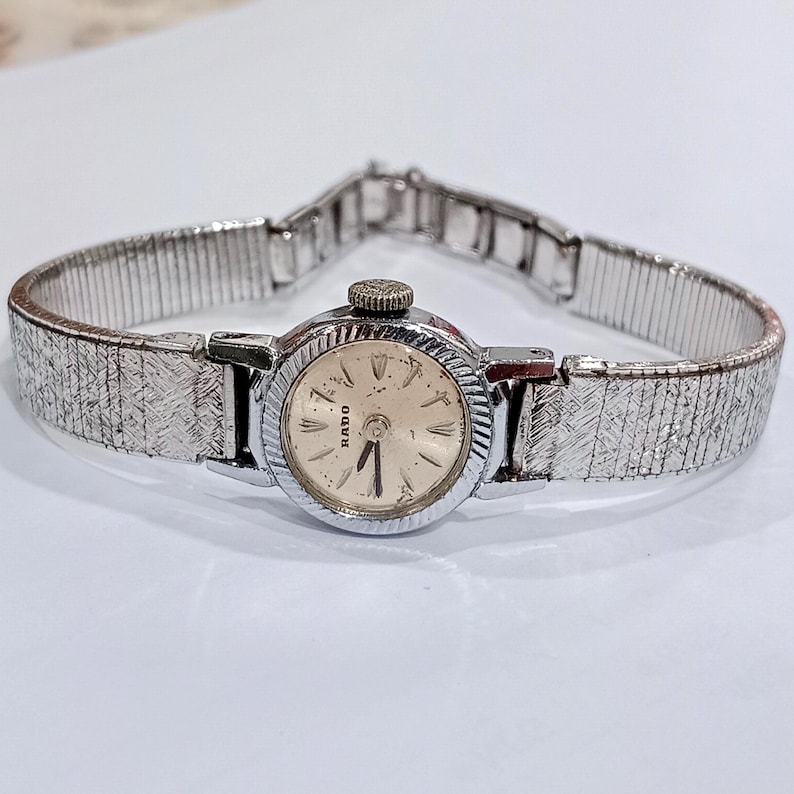Op de afbeelding: Een vintage Rado polshorloge met een zilverkleurige, getextureerde band en een ronde wijzerplaat. De wijzerplaat is cr&egrave;mekleurig met zwarte wijzers en uurmarkeringen. Het horloge heeft een kleine, getextureerde kroon en een sluiting.