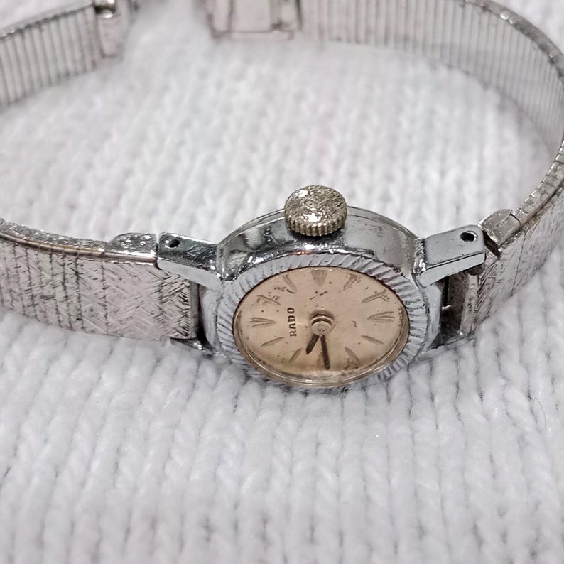 Op de afbeelding: Een vintage Rado polshorloge met een zilverkleurige kast en band. De wijzerplaat van het horloge is lichtgoudkleurig met zwarte wijzers en markeringen. De band heeft een textuurontwerp. Het horloge ligt op een wit, getextureerd oppervlak.