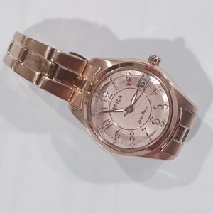 Puede incluir: Un reloj de pulsera Spica de energ&iacute;a solar, de color oro rosa, con una correa a juego. La esfera del reloj muestra la hora con n&uacute;meros ar&aacute;bigos y la fecha. El reloj est&aacute; en &aacute;ngulo, mostrando la correa y la esfera.