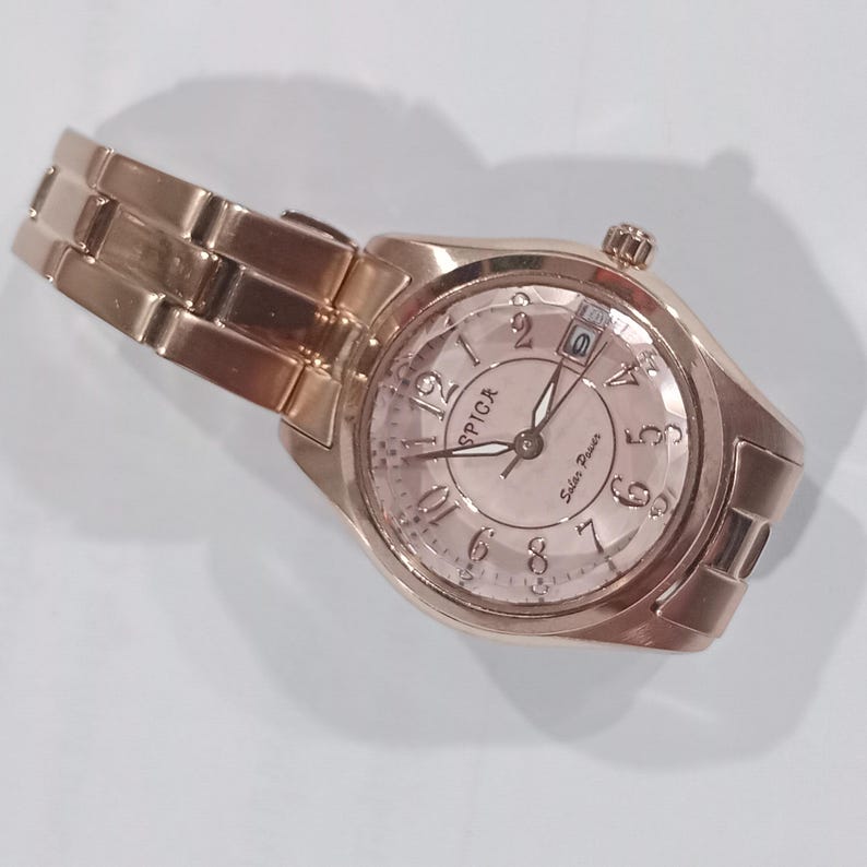 Puede incluir: Un reloj de pulsera Spica de energ&iacute;a solar, de color oro rosa, con una correa a juego. La esfera del reloj muestra la hora con n&uacute;meros ar&aacute;bigos y la fecha. El reloj est&aacute; en &aacute;ngulo, mostrando la correa y la esfera.