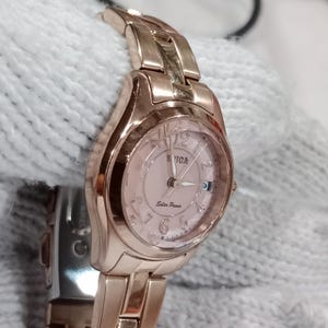 Puede incluir: Un reloj de pulsera en tono oro rosa con esfera rosa claro. El reloj muestra la marca "SIGA" y las palabras "Solar Power". El reloj tiene una pantalla de fecha y una correa de eslabones.