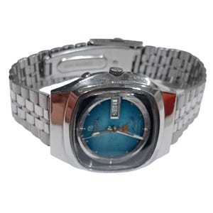Puede incluir: Un reloj de pulsera plateado con una esfera redonda de color azul brillante dentro de una caja cuadrada. El reloj incluye una correa de eslabones met&aacute;licos, manecillas plateadas y una ventana de d&iacute;a y fecha en la posici&oacute;n de las tres.