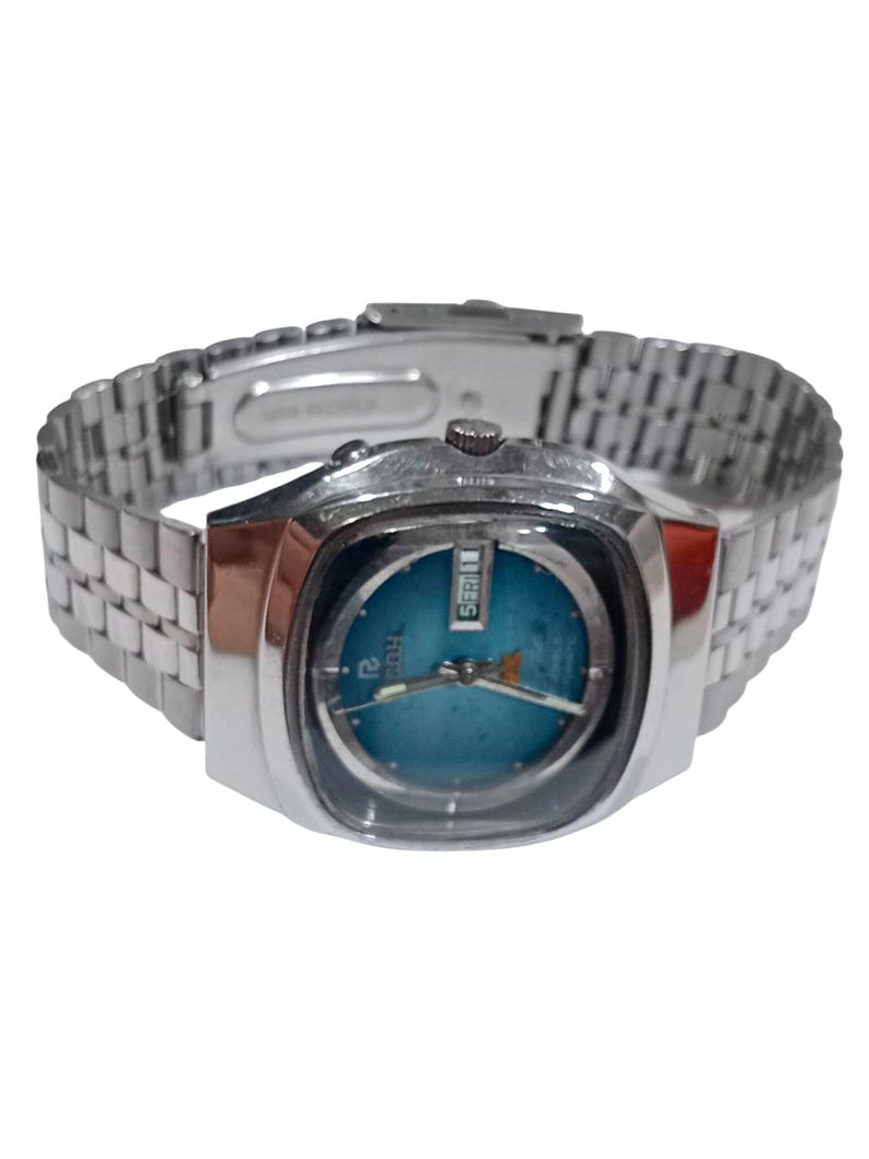Puede incluir: Un reloj de pulsera plateado con una esfera redonda de color azul brillante dentro de una caja cuadrada. El reloj incluye una correa de eslabones met&aacute;licos, manecillas plateadas y una ventana de d&iacute;a y fecha en la posici&oacute;n de las tres.