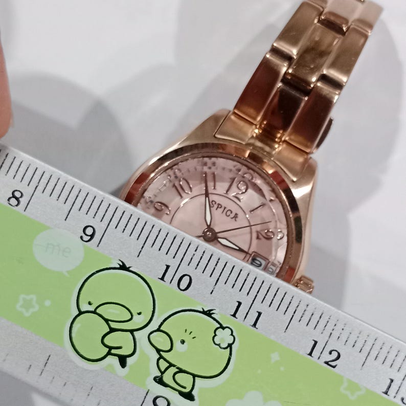 Puede incluir: Un reloj de pulsera SPICA en oro rosa con esfera rosa claro y correa a juego. La esfera del reloj muestra el nombre de la marca y la hora. El reloj se mide con una regla.