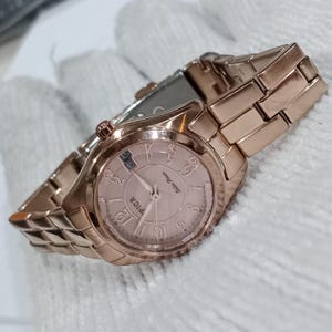 Puede incluir: Un reloj de pulsera de oro rosa con una correa a juego. La esfera del reloj es de color rosa claro con n&uacute;meros y agujas negros. El reloj tiene la palabra "PICA" impresa en la esfera.