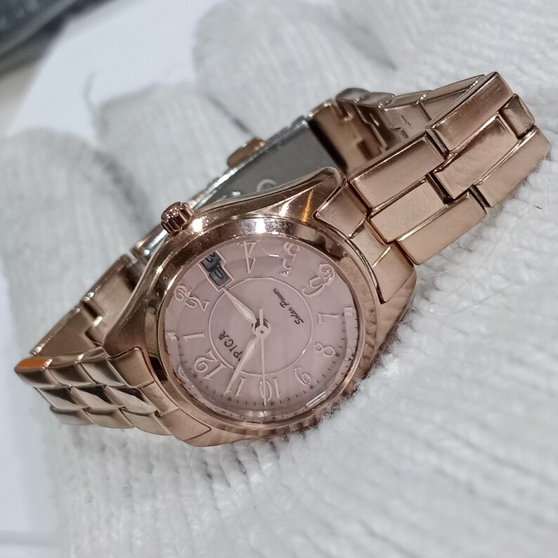 Puede incluir: Un reloj de pulsera de oro rosa con una correa a juego. La esfera del reloj es de color rosa claro con n&uacute;meros y agujas negros. El reloj tiene la palabra "PICA" impresa en la esfera.