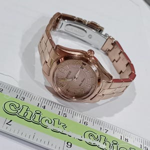 Puede incluir: Un reloj SPICA de oro rosa con una pulsera de eslabones a juego. La esfera del reloj es de color rosa claro con n&uacute;meros y agujas negros. El texto "SPICA" y "Solar Power" es visible.