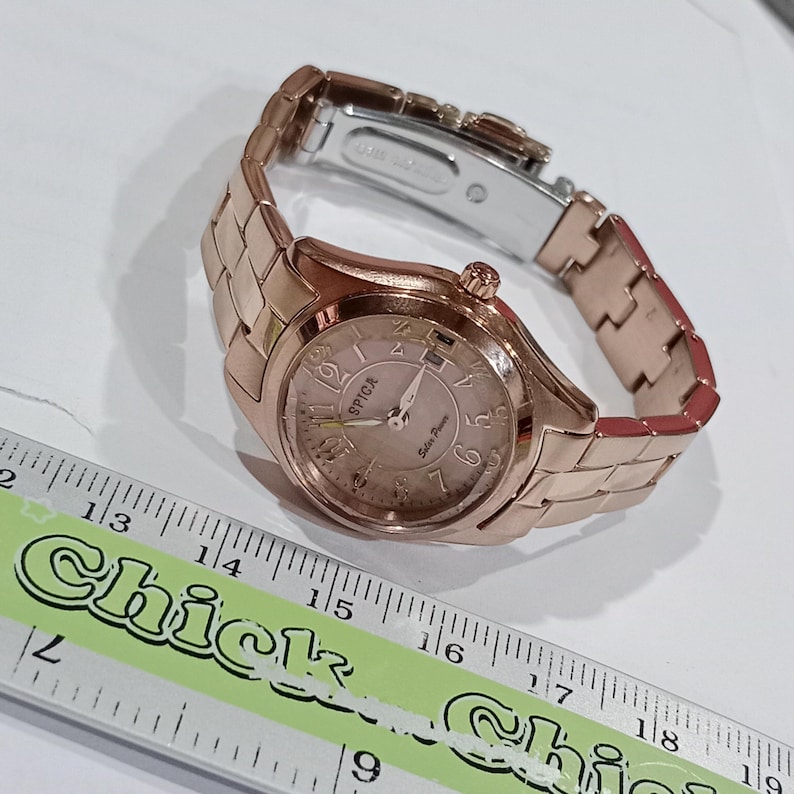 Puede incluir: Un reloj SPICA de oro rosa con una pulsera de eslabones a juego. La esfera del reloj es de color rosa claro con n&uacute;meros y agujas negros. El texto "SPICA" y "Solar Power" es visible.