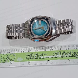 Puede incluir: Un reloj de pulsera autom&aacute;tico Ricoh plateado con esfera redonda color verde azulado, manecillas plateadas y ventana de d&iacute;a y fecha en la posici&oacute;n de las tres. El reloj tiene una correa de eslabones met&aacute;licos sobre una regla de 19 cm.