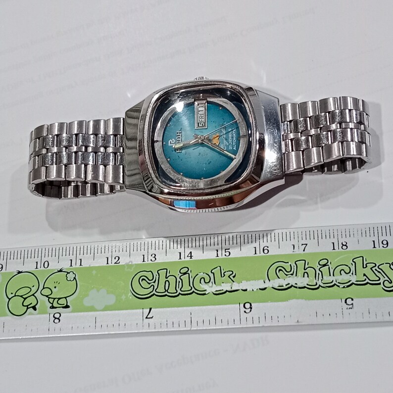 Puede incluir: Un reloj de pulsera autom&aacute;tico Ricoh plateado con esfera redonda color verde azulado, manecillas plateadas y ventana de d&iacute;a y fecha en la posici&oacute;n de las tres. El reloj tiene una correa de eslabones met&aacute;licos sobre una regla de 19 cm.