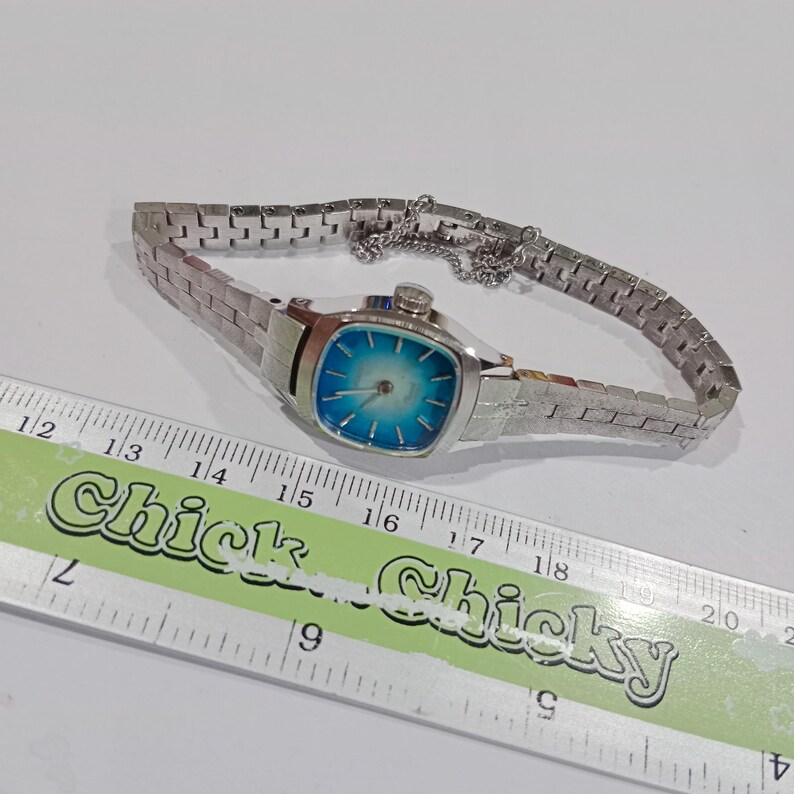 K&ouml;nnte beinhalten: Eine Vintage-Armbanduhr in Silberoptik mit quadratischem Zifferblatt und blau-wei&szlig;em Farbverlauf. Die Uhr hat ein Metallgliederarmband und eine kleine Kette. Ein Lineal befindet sich im Vordergrund.