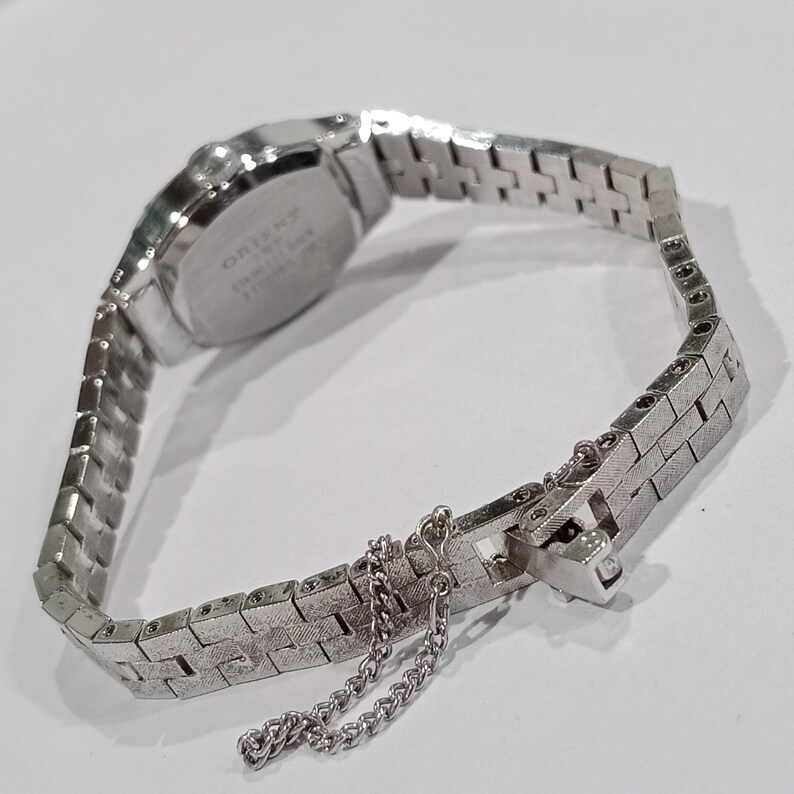 K&ouml;nnte beinhalten: Eine silberfarbene Orient-Armbanduhr mit rechteckigem Zifferblatt und strukturiertem Metallarmband. Das Armband ist mit einer Sicherheitskette verbunden. Die Uhr wird auf einer wei&szlig;en Oberfl&auml;che pr&auml;sentiert.