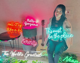 May include: A woman sits in front of a wall with neon signs that read "Hello Gorgeous", "Roll it up, take a hit", "Serenity", "This must be the place", and "The World's Greatest".