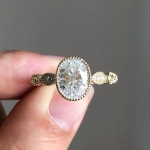 May include: A gold-toned ring featuring an oval-cut, clear gemstone as the centerpiece. The ring has an ornate, detailed band with smaller accent stones on either side of the main stone. The ring is held up by a person's fingers.