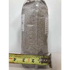 May include: A clear plastic bag filled with small, light-colored granules. A yellow measuring tape shows the bag is approximately 4 inches wide. The bag has a white label with text. The product appears to be a gardening or landscaping material.