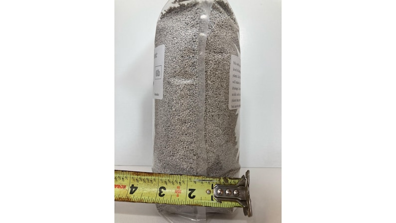May include: A clear plastic bag filled with small, light-colored granules. A yellow measuring tape shows the bag is approximately 4 inches wide. The bag has a white label with text. The product appears to be a gardening or landscaping material.