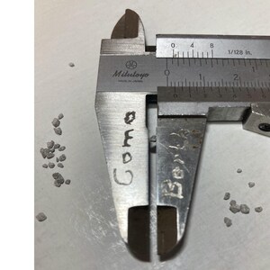 May include: A close-up of a Mitutoyo caliper measuring a small, dark-colored object. The caliper is silver with black markings, and the object is a small, irregular shape. The word "Cora" is written on the caliper.