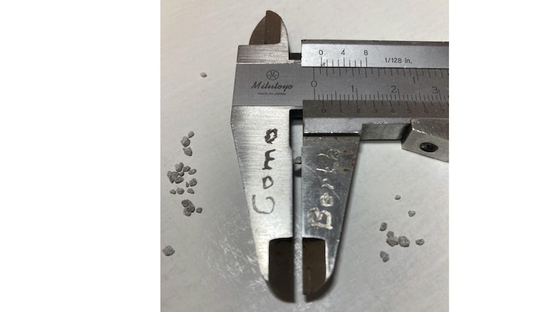 May include: A close-up of a Mitutoyo caliper measuring a small, dark-colored object. The caliper is silver with black markings, and the object is a small, irregular shape. The word "Cora" is written on the caliper.