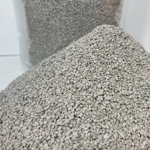 May include: A close-up shot of a pile of small, light gray, granular material. The material is in focus, with a clear plastic bag filled with the same material visible in the background. The granules appear to be uniform in size and shape.