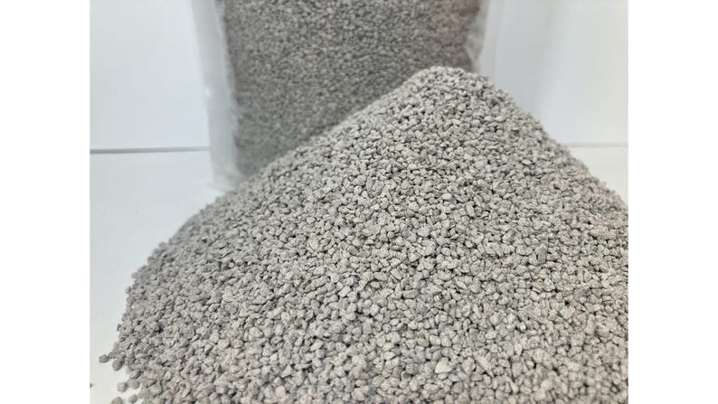 May include: A close-up shot of a pile of small, light gray, granular material. The material is in focus, with a clear plastic bag filled with the same material visible in the background. The granules appear to be uniform in size and shape.