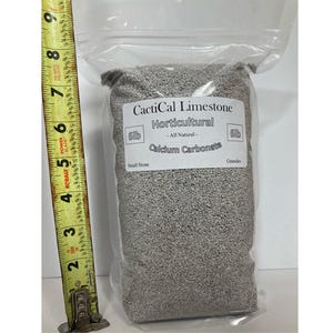 May include: A clear plastic bag filled with gray horticultural limestone. The bag has a white label with the text "CactiCal Limestone Horticultural All Natural Calcium Carbonate". A yellow tape measure is positioned next to the bag.