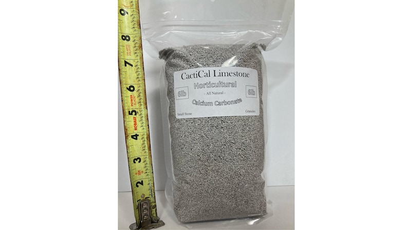 May include: A clear plastic bag filled with gray horticultural limestone. The bag has a white label with the text "CactiCal Limestone Horticultural All Natural Calcium Carbonate". A yellow tape measure is positioned next to the bag.