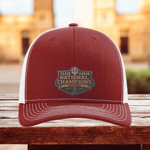 May include: A maroon and white trucker hat with the text "NATIONAL CHAMPIONS HOOSIERS 2025" in a shield design. The hat has a curved brim and a mesh back. The hat is sitting on a wooden surface.