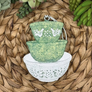 May include: A decorative ornament featuring three stacked ceramic bowls. The top two bowls are green with white bird and floral designs. The bottom bowl is white with a detailed lace-like pattern. The ornament hangs from a white string.