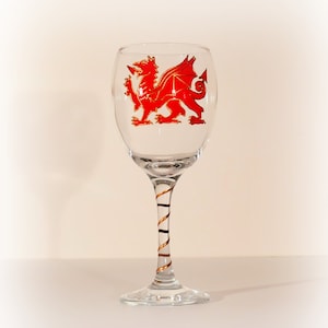 May include: A clear glass wine glass with a red dragon design on the front and a gold and black swirl design on the stem.