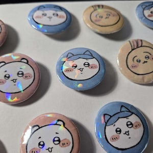 以下が含まれることがあります： ホログラフィックアクセントが付いた、丸い漫画スタイルのボタンのコレクション。ボタンには、ピンク、青、タンなど、さまざまな色のキュートな動物の顔が描かれています。デザインは、日本の文化で人気の高い、かわいい美学を持っています。
