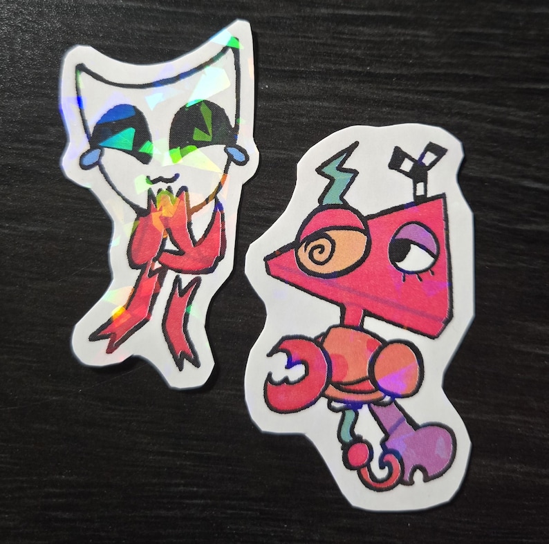 May include: Two holographic stickers featuring cartoon characters. One is a white cat-like figure with a red scarf, the other is a red robot with a spiral eye and crab-like claws. Both stickers have a colorful, iridescent sheen.