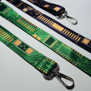 Puede incluir: Dos cordones de tela con un patrón de placa de circuito impreso. Uno es verde con líneas y componentes dorados, mientras que el otro es negro con detalles dorados. Ambos incluyen un mosquetón de metal en tono plateado.