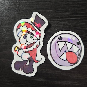 May include: Two holographic stickers. One features a cartoon character in a red and white outfit with a top hat. The other is a purple, round character with a large mouth and teeth.