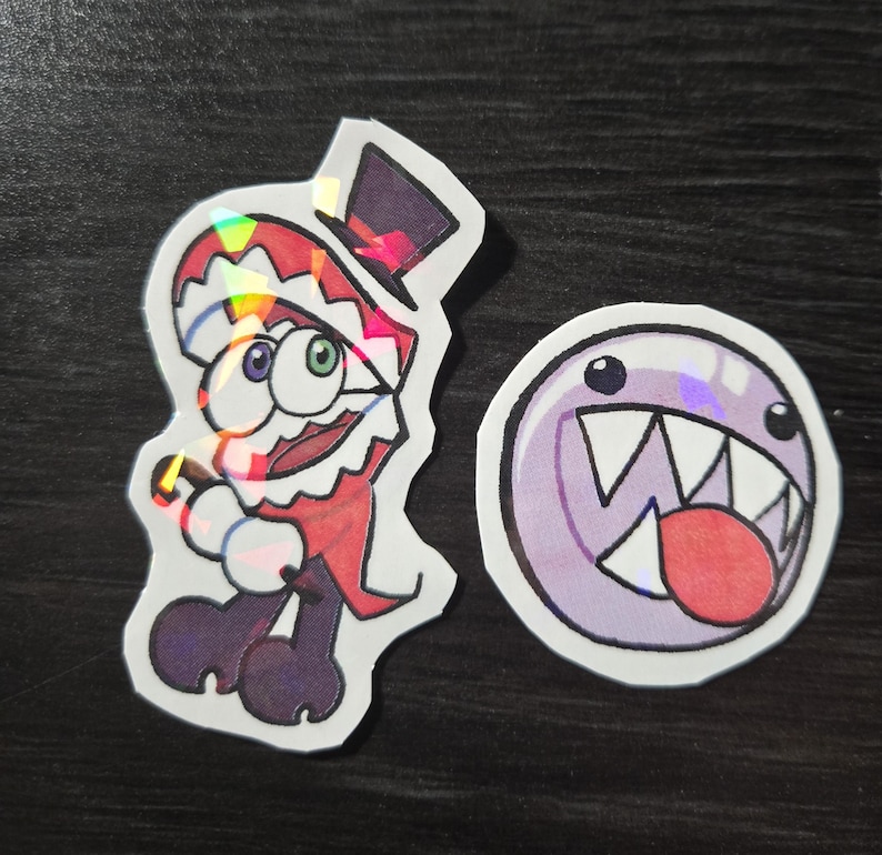 May include: Two holographic stickers. One features a cartoon character in a red and white outfit with a top hat. The other is a purple, round character with a large mouth and teeth.