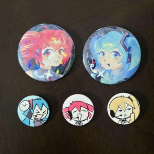 May include: Two large circular holographic pinback buttons featuring anime-style characters with pink and blue hair, alongside three smaller circular buttons with simplified black-and-white line art illustrations of characters wearing headsets.
