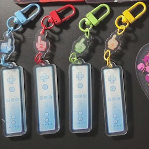 May include: Four acrylic keychains shaped like game controllers, each with a matching colored lobster clasp and a small charm featuring a numbered pointing finger. The controllers are light blue with white buttons and numbered 1, 2, 3, and 4.