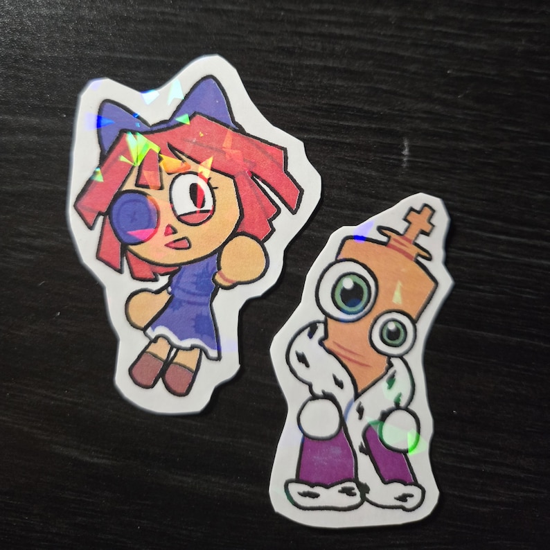 May include: Two colorful stickers featuring cartoon characters. One sticker depicts a girl with red hair, a blue dress, and a bow. The other sticker shows a king with a crown and a regal robe. Both stickers have a holographic effect.